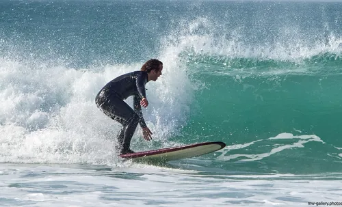 This landscape photograph was captured on a spring afternoon and features a Cornish surfer riding a wave near the coast at Sennen Cove in Cornwall, United Kingdom. The image shows the surfer, dressed in a wetsuit, skilfully balancing on a surfboard as waves break behind, with the turquoise water and white foam characteristic of this nature-rich beach environment. The ocean is lively with rolling waves, highlighting the popularity of Sennen Cove among surfers and beach visitors in Cornwall. The scene is set along the scenic coast, typical of the rugged beauty found in the United Kingdom, with the natural elements of the beach and the dynamic action of the surfers central to the composition.